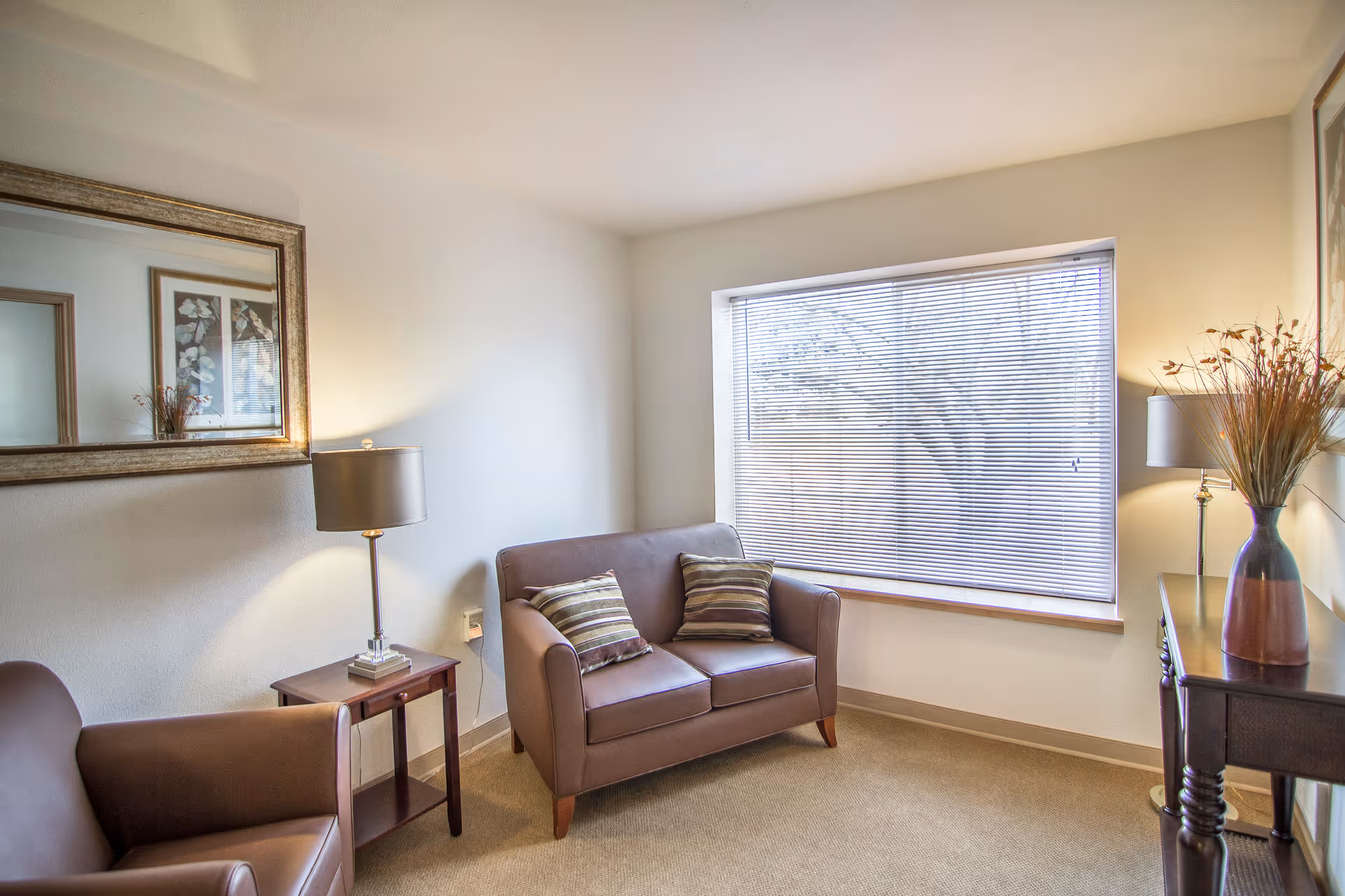 A cozy living room area with a brown loveseat and armchair, both with striped cushions. There is a wooden side table with a lamp between the chairs, a large window with blinds letting in natural light, a decorative vase with dried plants on a wooden console table, and a large framed mirror on the wall.