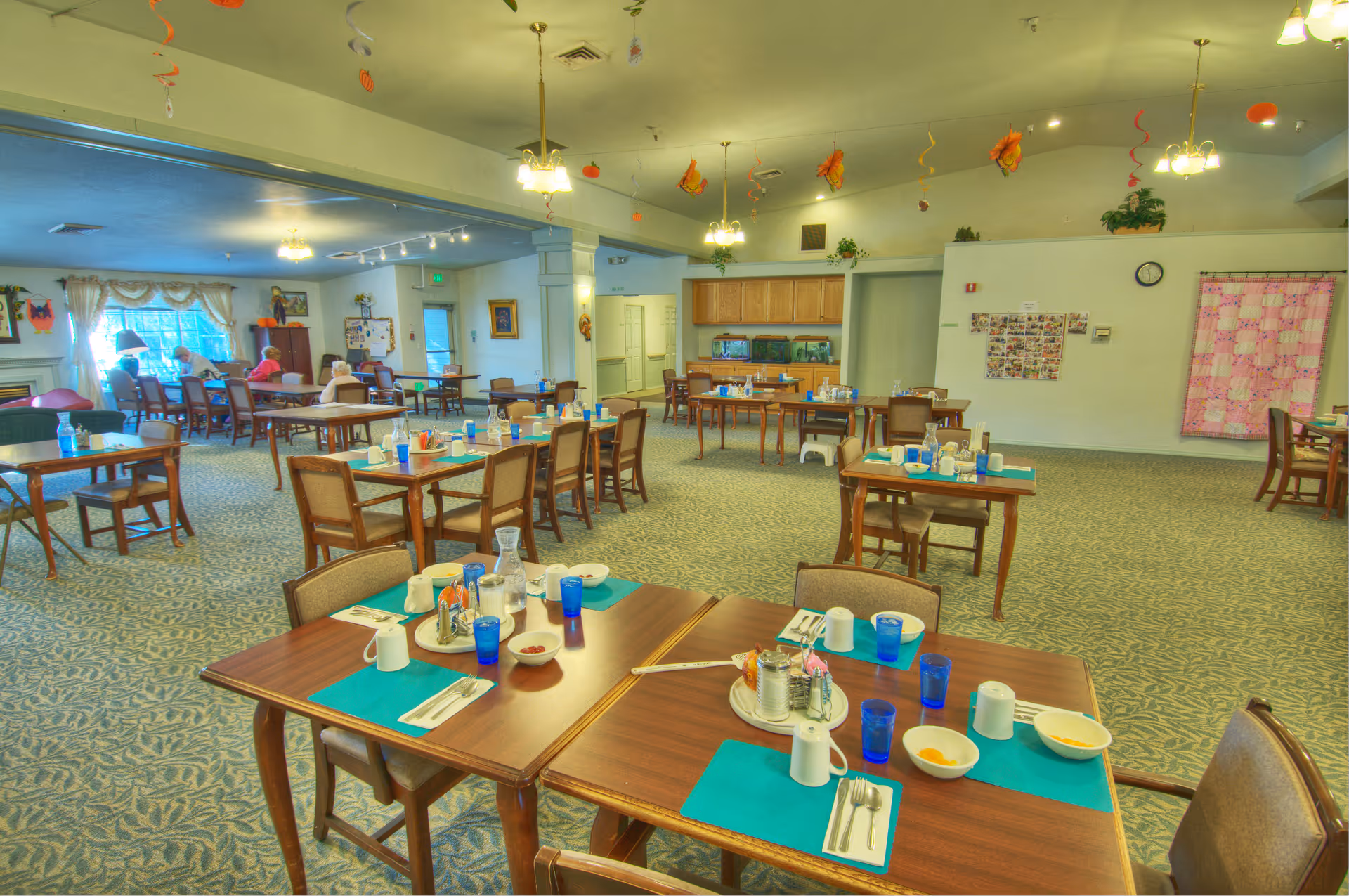 Spacious assisted living dining room with multiple set tables, teal placemats and blue glasses under hanging lights and ceiling decorations.