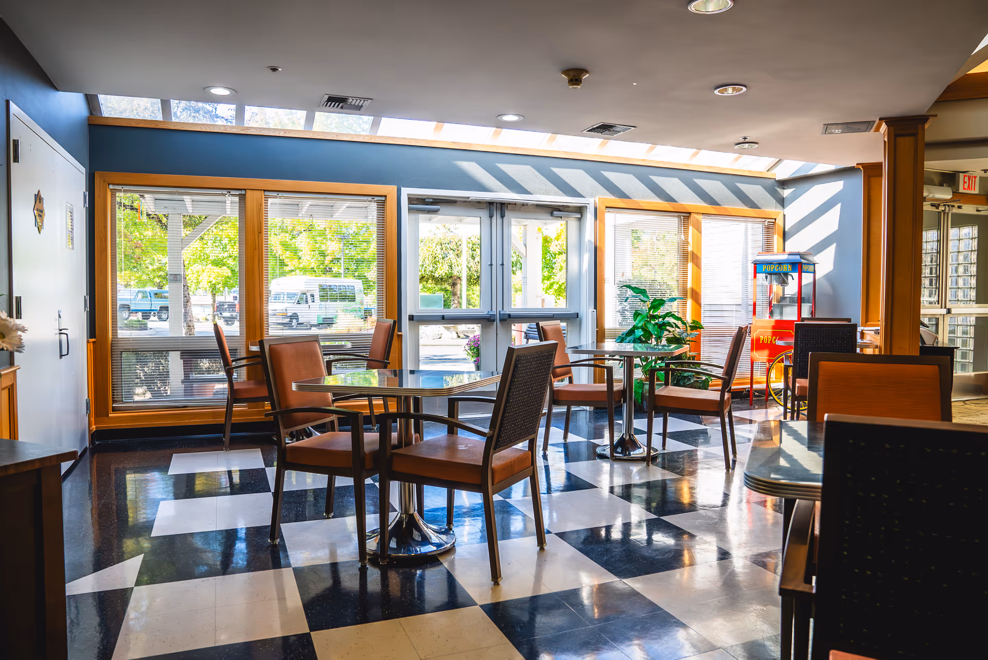 Bright common area with checkered black and white floor tiles, several round tables with brown chairs, large windows letting in natural light, a popcorn machine in the corner, and a glass door leading outside.