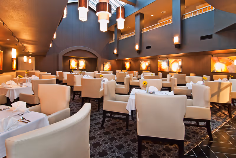 A spacious, elegant dining room with multiple tables covered in white tablecloths, each set with napkins, cutlery, and a single yellow flower in a vase. The room features beige upholstered chairs, warm lighting from modern hanging fixtures, and colorful abstract artwork on the walls. There is a high ceiling with a skylight and a second-floor balcony overlooking the dining area.