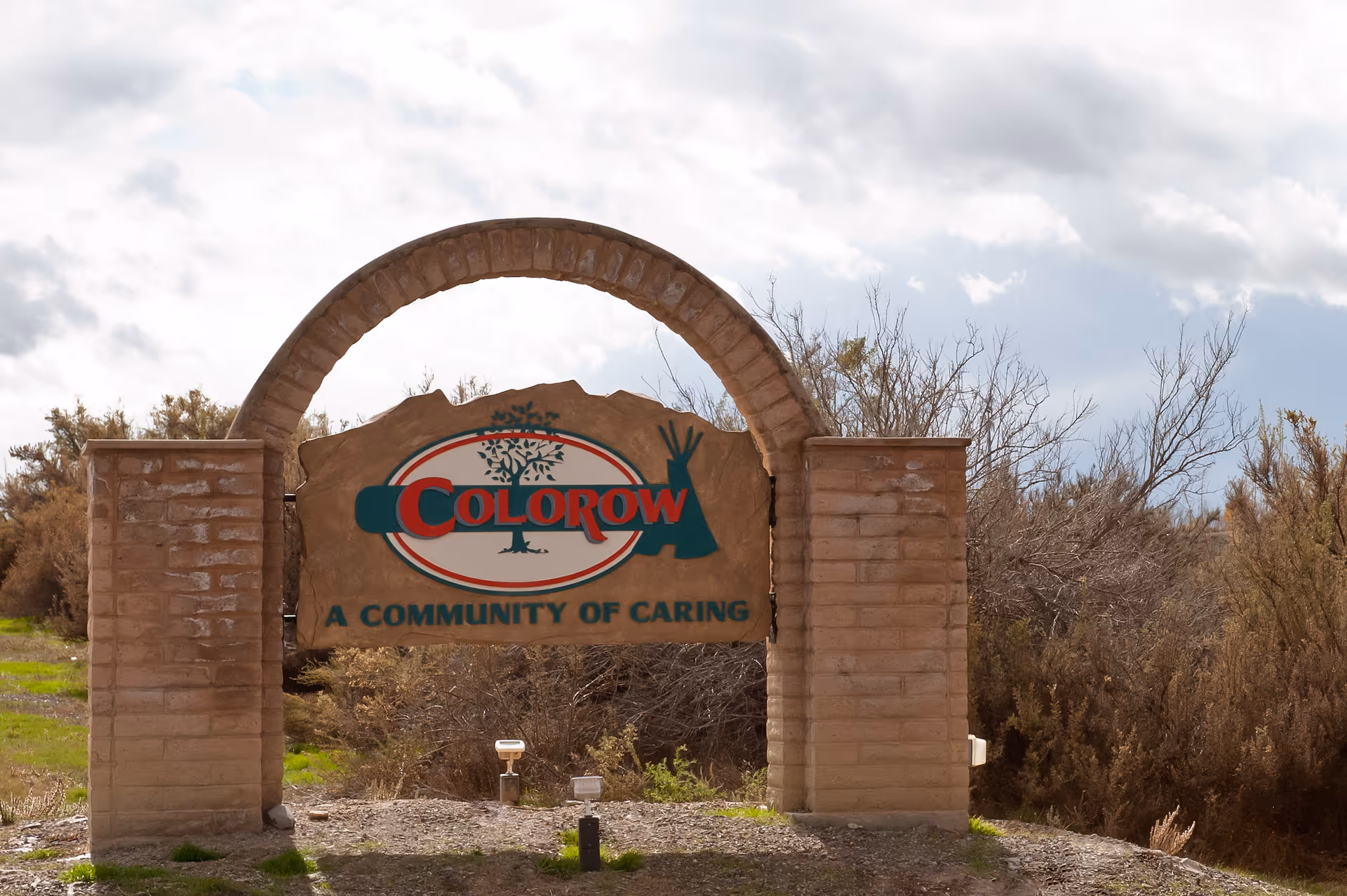 Stone archway sign for Colorow Care Center with the text 'A Community of Caring' surrounded by natural shrubbery and cloudy sky in the background.