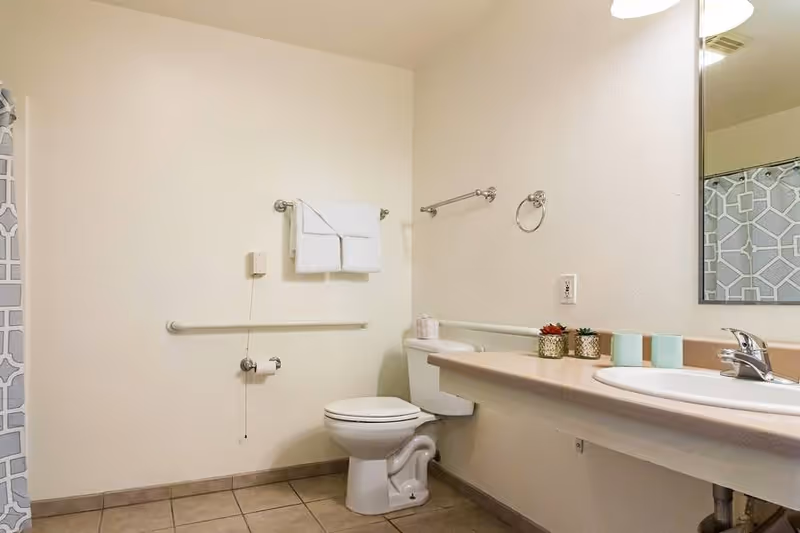 A clean and accessible bathroom with a toilet, a towel rack with folded white towels, a grab bar, a sink with a countertop, a mirror, and a shower curtain with a geometric pattern.
