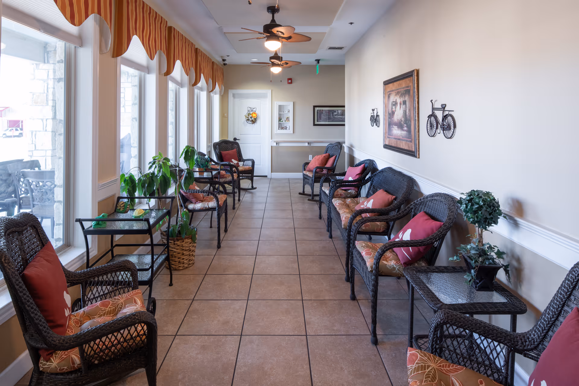 A bright, narrow seating area with large windows on one side and a row of wicker chairs with cushions and pillows along the opposite wall. Small tables with plants and decorative items are placed between the chairs. Ceiling fans with lights hang from the ceiling, and the walls are decorated with framed artwork and metal bicycle sculptures.