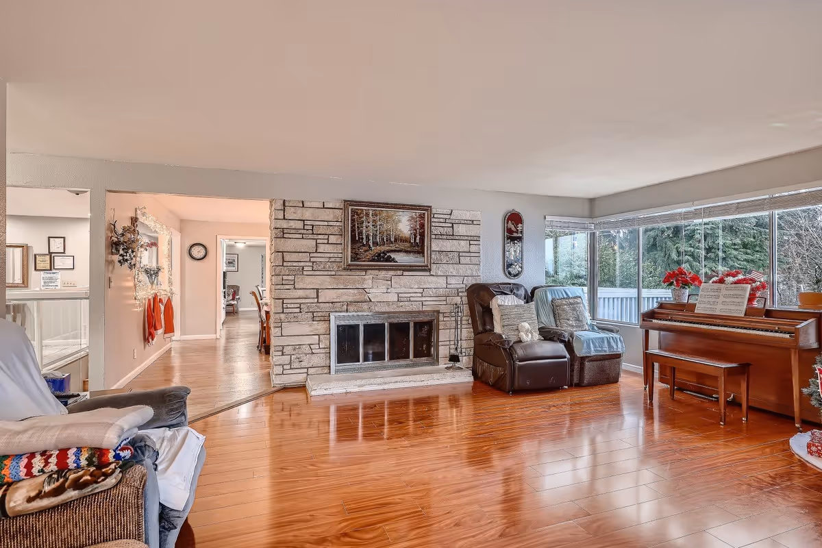 A cozy living room with polished wooden floors, a stone fireplace with a framed painting above it, two recliner chairs with cushions and throws, a wooden piano with sheet music and red poinsettia plants on top, and large windows showing green trees outside.