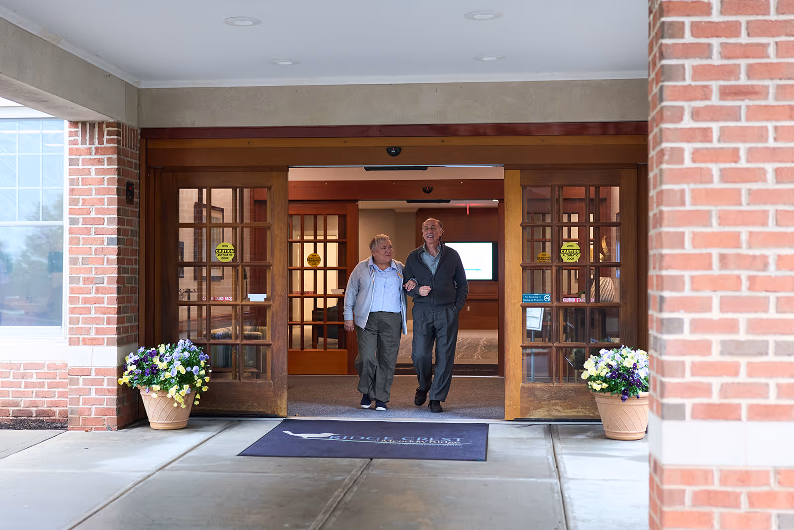 Two older adults walking arm-in-arm through the main entrance of a senior living facility flanked by potted flowers.