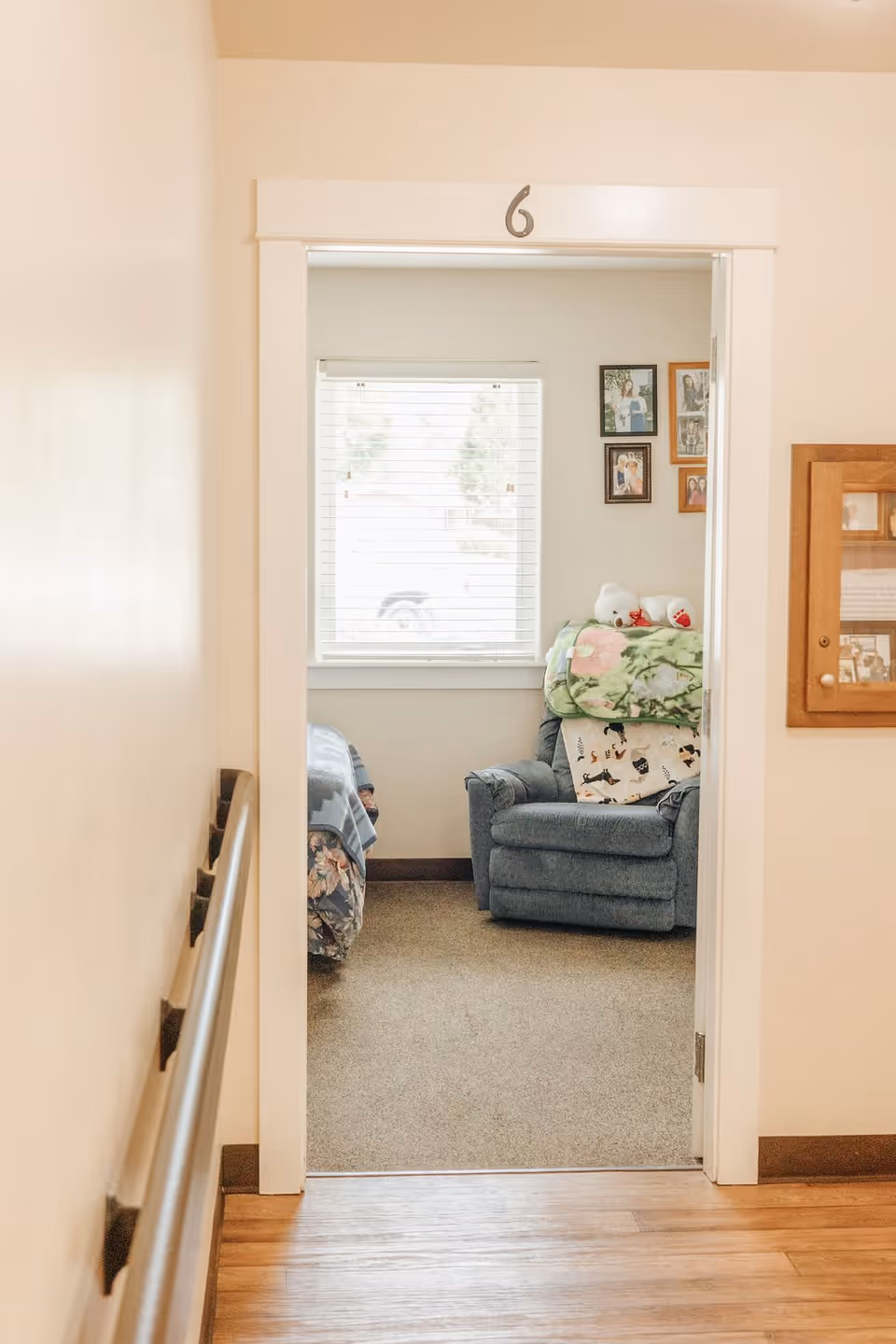 View through a doorway labeled with the number 6 into a cozy room with a window covered by blinds, a blue armchair with a floral blanket and a pillow, and part of a bed with a patterned blanket. The walls are light-colored and there is a wooden floor in the hallway outside the room.