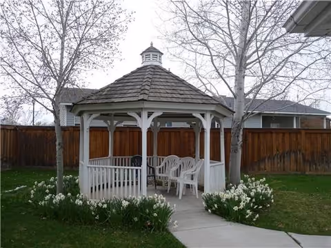 White wooden gazebo with a shingled roof situated in a grassy yard with a concrete pathway leading to it. The gazebo contains several white plastic chairs and is surrounded by blooming white flowers and two leafless trees. A wooden fence and neighboring houses are visible in the background under a cloudy sky.
