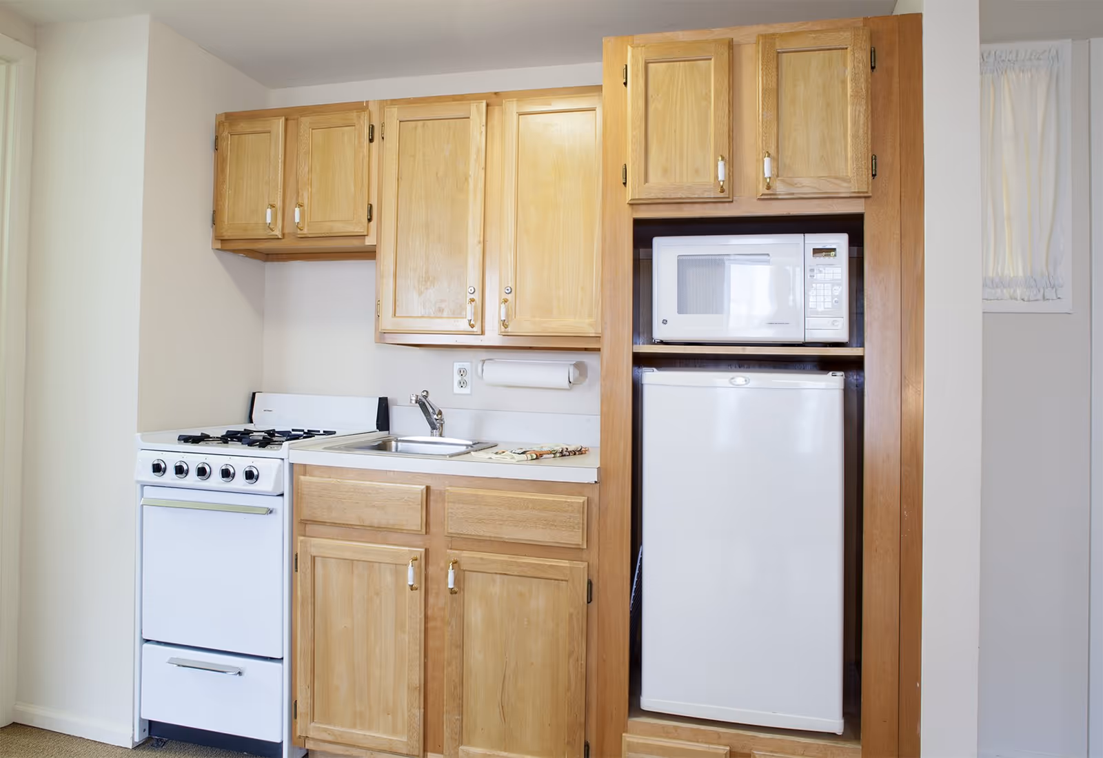 Small kitchen area with light wood cabinets, a white gas stove, a stainless steel sink, a white microwave, and a white mini refrigerator. There is a small window with white curtains on the right side.