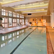 Indoor swimming pool with lounge chairs, large windows, and a high beamed ceiling reflected in the water.