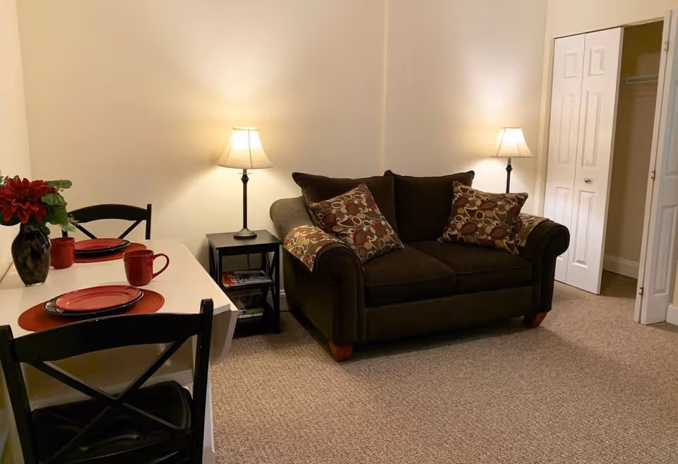 A cozy living area with a dark brown loveseat adorned with patterned cushions, two lit table lamps on either side, a small black side table with magazines, and a white dining table set with red plates, mugs, and a vase with red flowers. There is a closet with white bi-fold doors in the background.