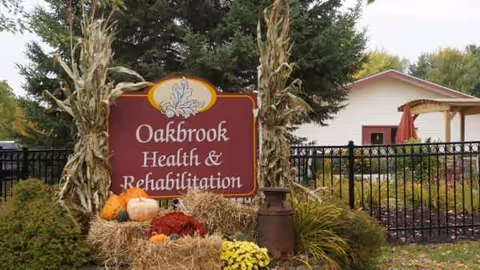 Oakbrook Health & Rehabilitation sign with autumn decorations outdoors near a fence.
