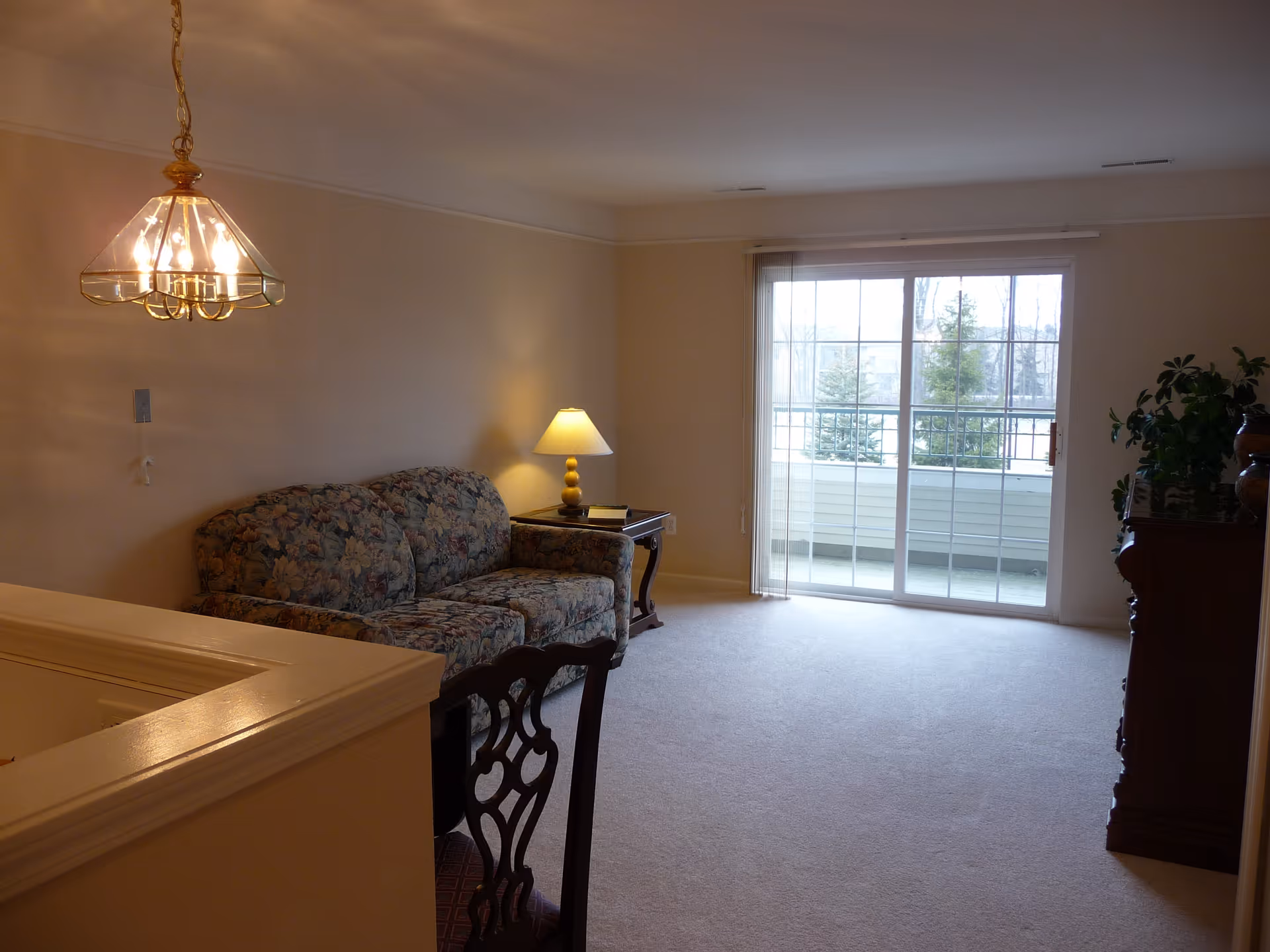 A living room with a floral patterned sofa, a wooden side table with a lamp, a wooden chair with a decorative backrest, a chandelier hanging from the ceiling, and a large sliding glass door leading to a balcony with a view of trees outside.