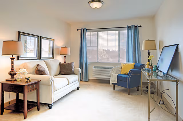 Bright furnished living room with a cream sofa, blue armchair, side tables, lamps and a TV console in front of a large window with blue curtains.