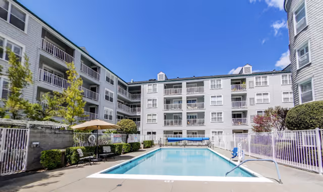 Outdoor swimming pool area surrounded by a multi-story residential building with balconies. There are benches, umbrellas, and greenery around the pool, with a clear blue sky above.