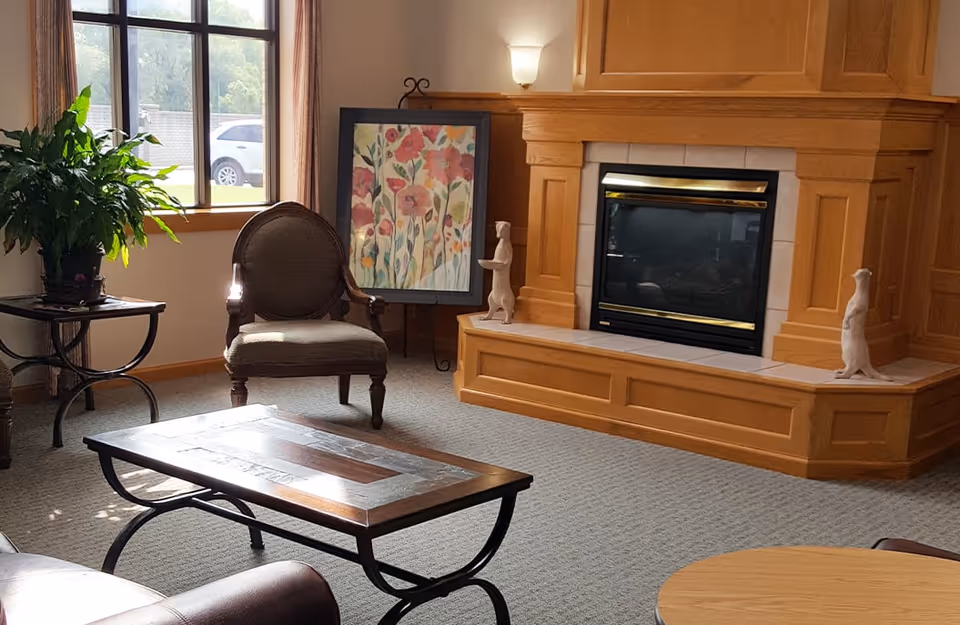 A cozy living room area featuring a wooden fireplace with tile accents, two small white animal figurines on the hearth, a framed floral painting resting on an easel, a cushioned armchair, a wooden coffee table with metal legs, a side table with a green potted plant, and a window with beige curtains letting in natural light.