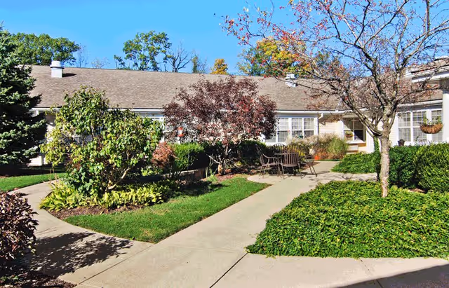 Outdoor courtyard area at Ontario Estates Senior Living with paved walkways, green grass, various shrubs and trees, and a seating area with a table and chairs under a clear blue sky.