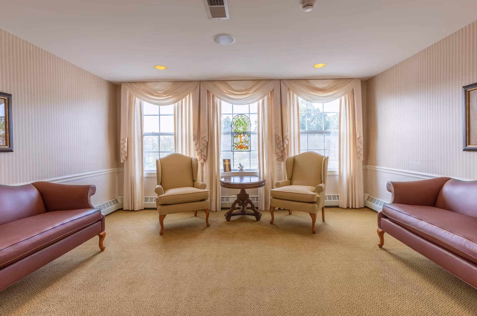 A cozy sitting area with two beige armchairs and a round wooden table between them, positioned in front of three large windows with cream-colored curtains. On either side of the room are two maroon leather sofas. The walls have light striped wallpaper and framed artwork, and the floor is carpeted in a neutral tone.