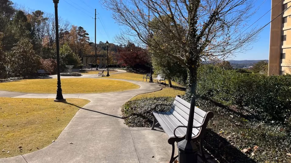 A sunny outdoor garden area with a winding concrete pathway, benches along the path, lamp posts, trees with some autumn foliage, and a building partially visible on the right side.