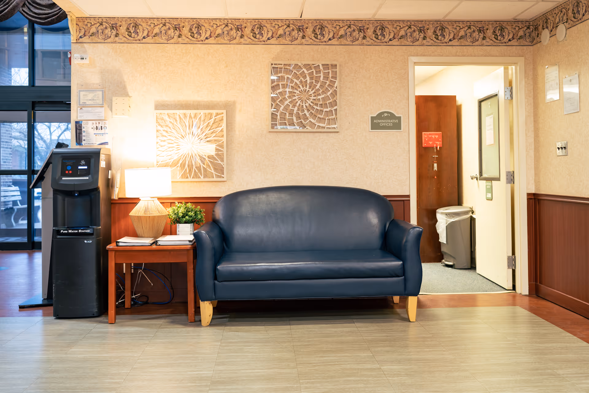 A waiting area with a dark blue leather loveseat, a wooden side table with a lamp and a small plant, and a black water dispenser. The wall behind has two decorative framed artworks and a sign indicating 'Administrative Offices' above an open door leading to an office space with a trash can visible inside.