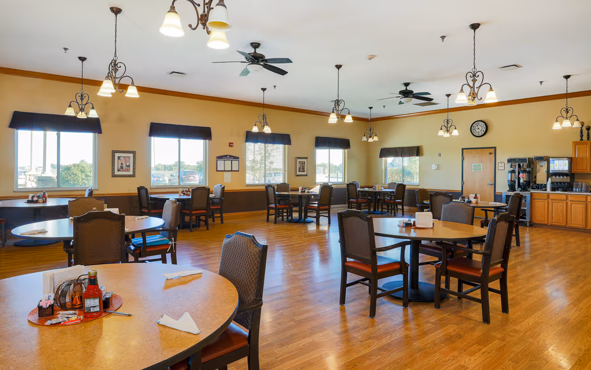 A spacious dining room with multiple round tables and chairs arranged neatly. The room has large windows with dark valances allowing natural light to fill the space. The walls are painted a warm yellow with wooden trim, and the floor is a polished wood. Ceiling fans and hanging light fixtures provide lighting and ventilation. In the back corner, there is a beverage station with coffee machines and a door. The room appears clean and inviting.