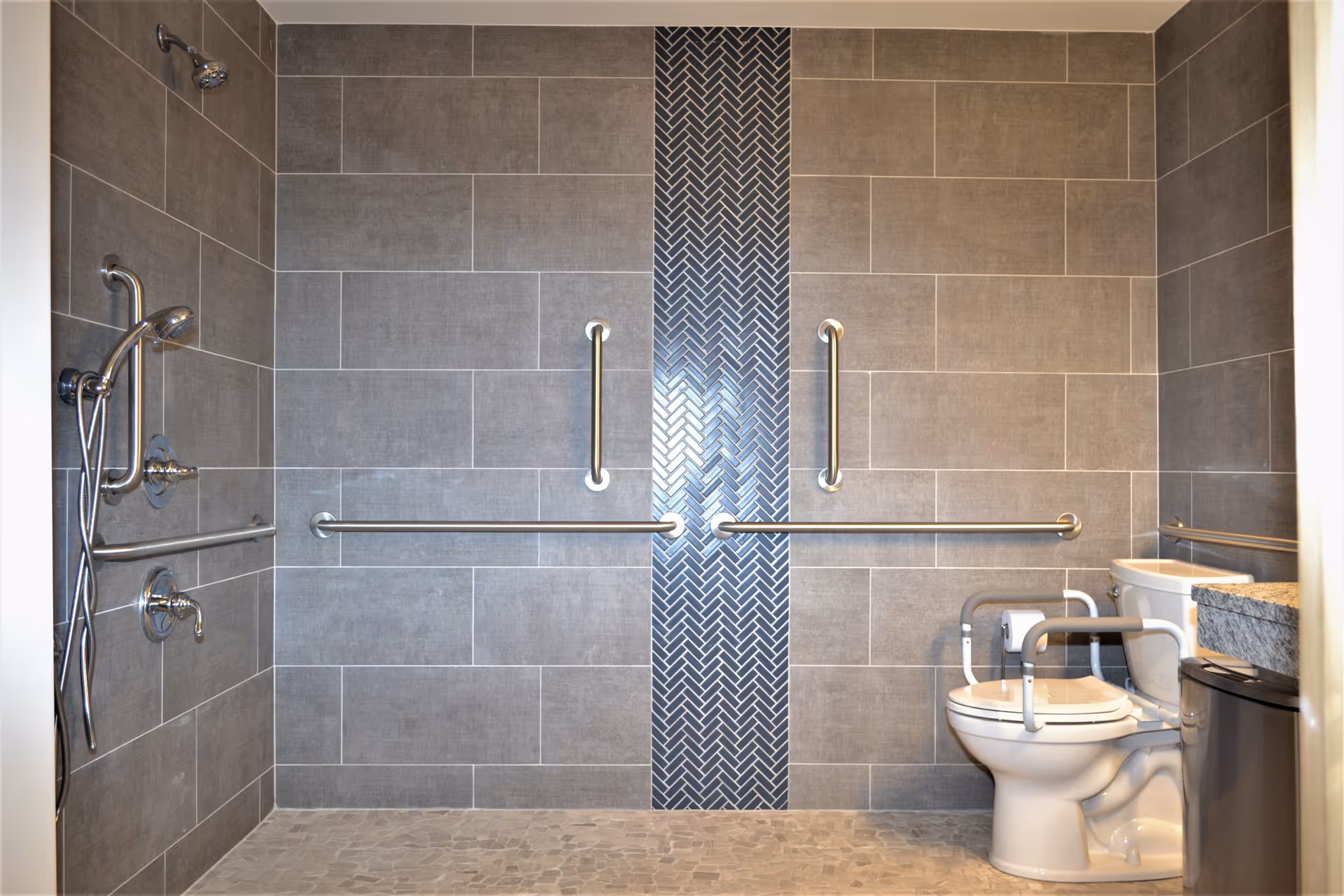 Accessible bathroom with gray tiled walls and floor, featuring multiple grab bars around a toilet and a walk-in shower area with a handheld showerhead and fixed showerhead.