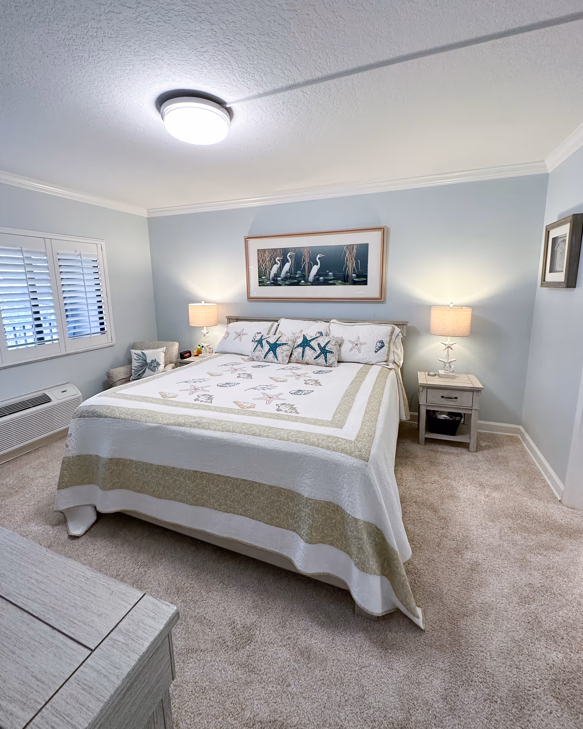 Bright bedroom with a large bed dressed in coastal-themed bedding, two bedside tables with lamps, a painting above the headboard and a shuttered window.