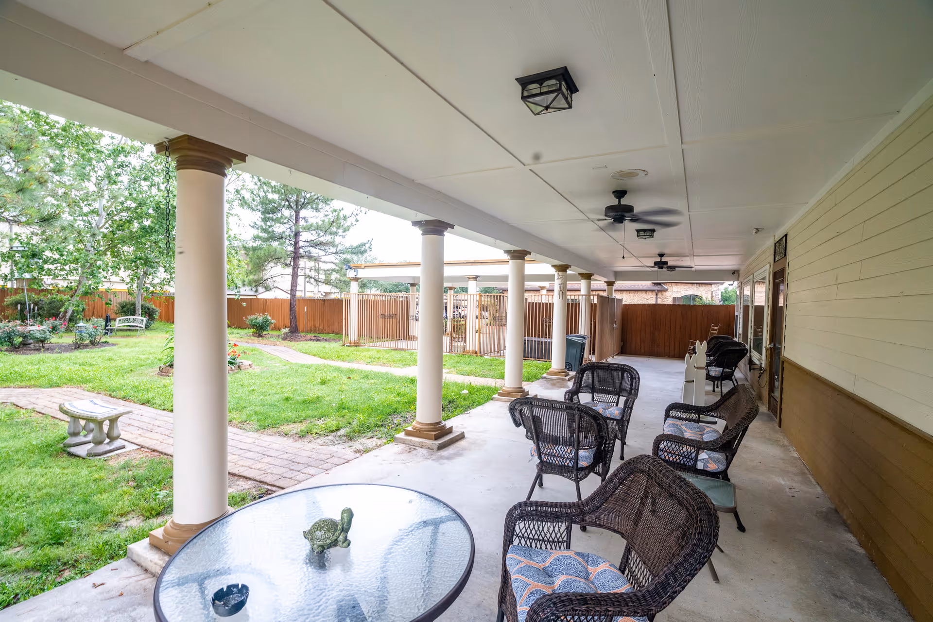 Covered outdoor patio area with several wicker chairs and a glass-top table with a small turtle decoration. The patio overlooks a grassy garden with trees, a swing, and a stone bench. Ceiling fans and light fixtures are mounted on the patio ceiling.