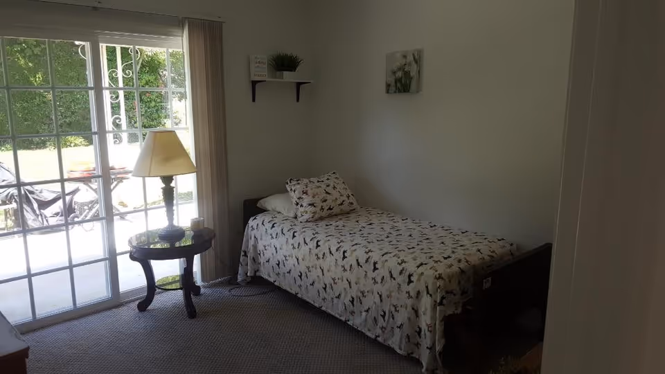 Small bedroom with a single bed covered in patterned bedding, a round side table with a lamp, and sliding glass doors to an outdoor patio.