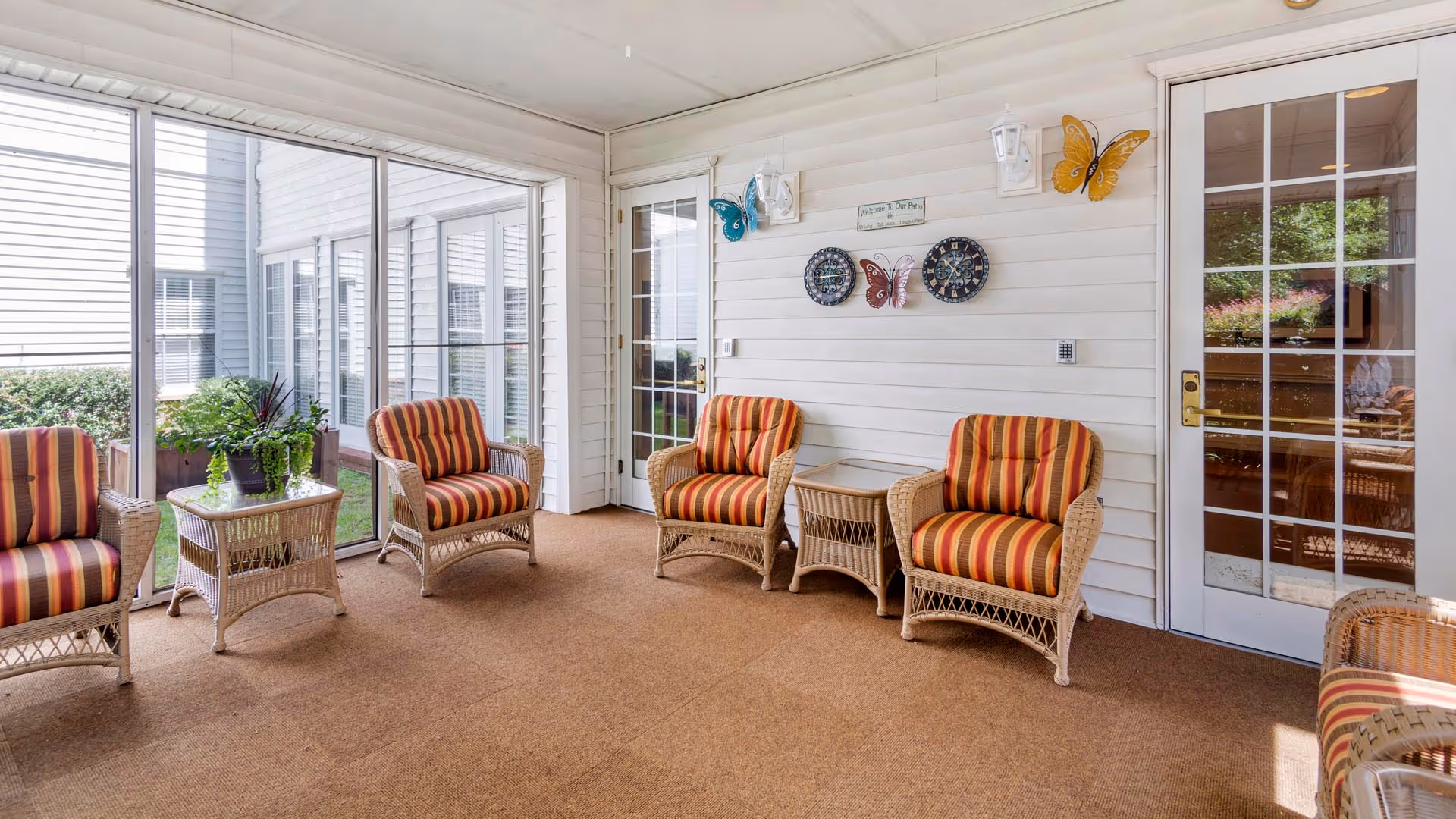 A bright enclosed patio area with four wicker armchairs featuring orange and red striped cushions, two wicker side tables with glass tops, and decorative butterfly wall hangings. Large windows and glass doors allow natural light to fill the space, and there are plants visible outside.