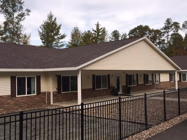Exterior view of a single-story assisted living facility building with beige siding, brown brick accents, and a dark shingled roof. The building has several windows with dark shutters and a covered entrance area. In front of the building is a black metal fence enclosing a gravel area. Tall trees are visible in the background under a cloudy sky.