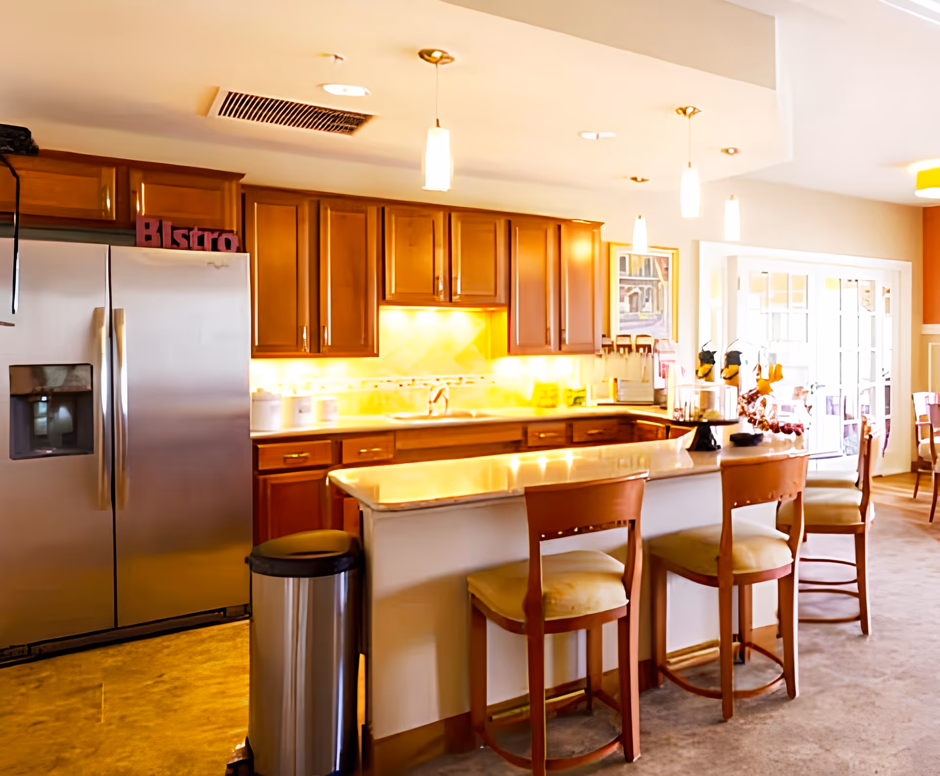 A bright kitchen area with wooden cabinets, a stainless steel refrigerator, a countertop with three wooden chairs, and pendant lights hanging from the ceiling. There is a trash can next to the counter and a sign that reads 'Bistro' on top of the refrigerator.