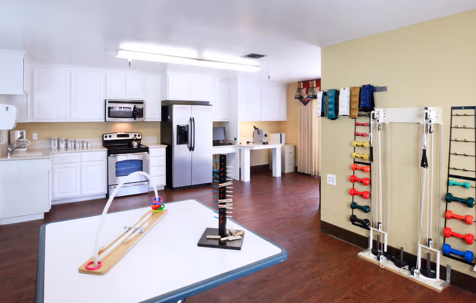 A bright room featuring a kitchen area with white cabinets, a stainless steel refrigerator, stove, and microwave. The room also contains a table with various therapy or activity tools, and a wall-mounted rack holding colorful dumbbells and exercise equipment, suggesting a multipurpose space for cooking and physical therapy or activities.