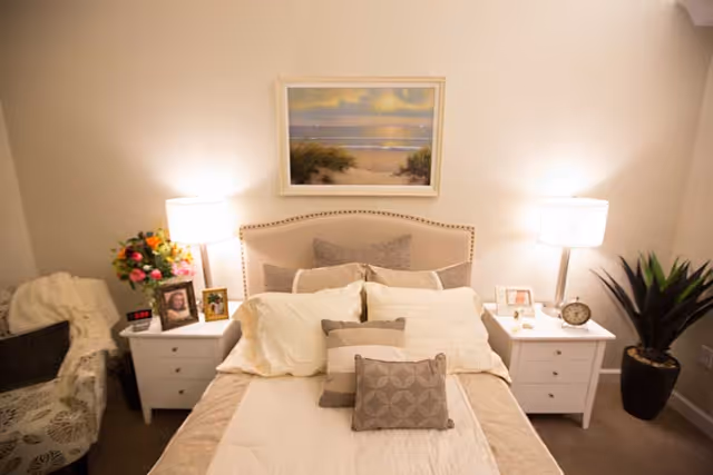 A cozy bedroom with a neatly made bed featuring multiple pillows and a beige headboard. On either side of the bed are white nightstands with lamps, framed photos, and decorative items. A floral armchair with a white throw blanket is positioned to the left, and a potted plant is on the right. Above the bed hangs a framed painting of a beach scene at sunset.