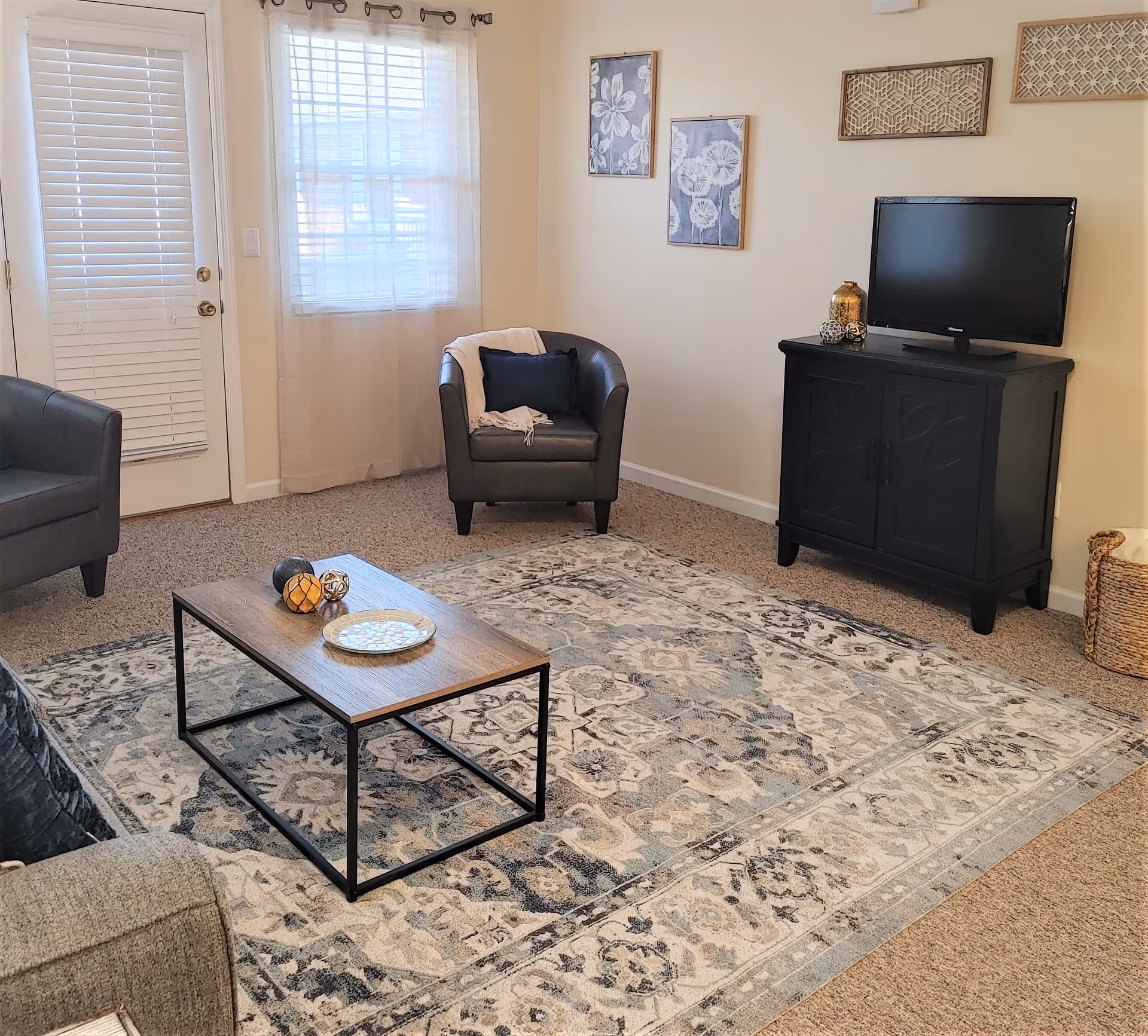 A cozy living room with a patterned area rug, a wooden coffee table with decorative items, two black armchairs, a TV on a black cabinet, and wall art. There is a window with sheer curtains and a door with blinds.