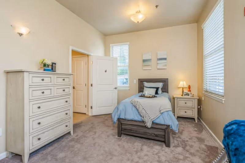A cozy bedroom with a single bed dressed in blue bedding and a gray throw blanket. The room features beige walls, carpeted floor, a wooden dresser with books and flowers on top, a nightstand with a lamp and picture frame, two windows with blinds, and two small paintings above the bed.