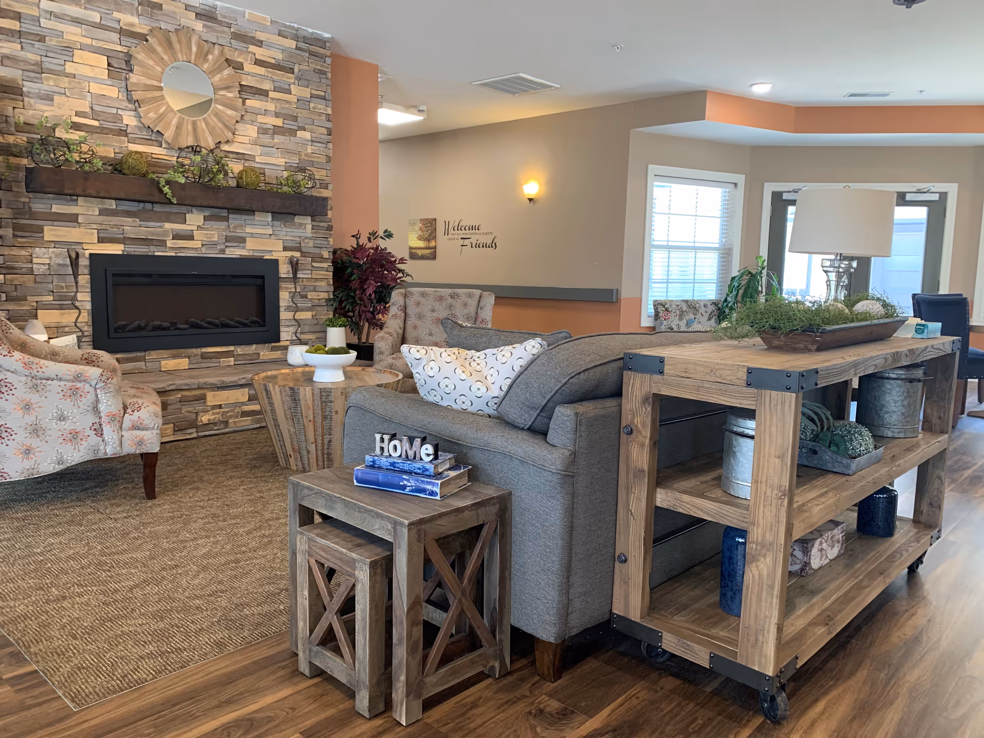 A cozy living room area in a retirement village featuring a stone fireplace with a decorative round mirror above it, two floral armchairs, a gray sofa with patterned pillows, a wooden side table with books and a 'Home' decoration, and a wooden shelving unit with plants and decorative items. The room has wooden flooring and a welcoming ambiance.