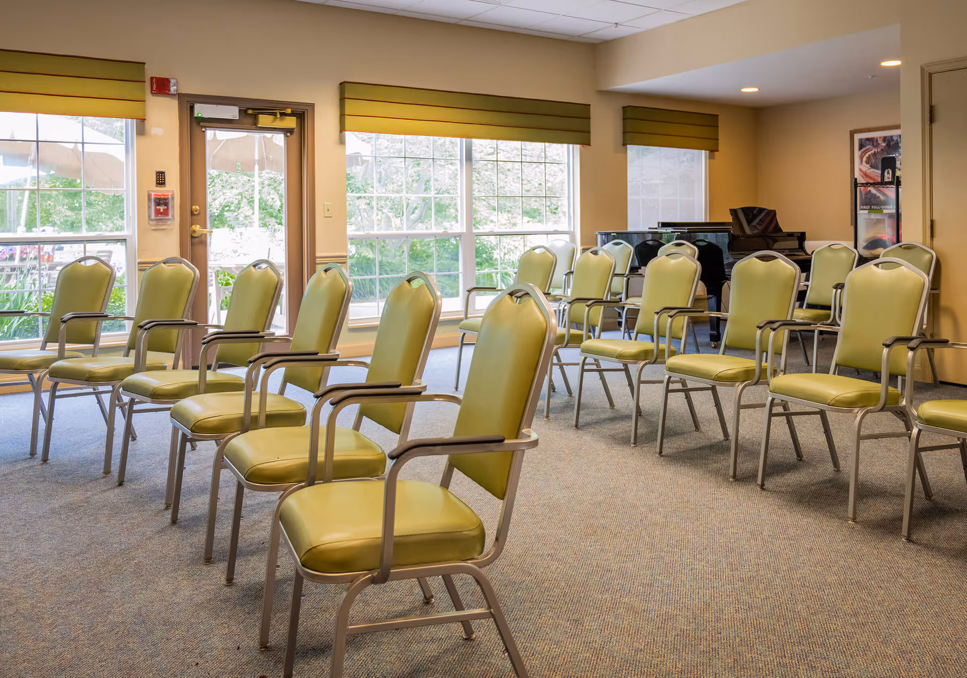 A room with multiple green cushioned chairs arranged in rows facing a black grand piano near the back wall. Large windows with green valances let in natural light, and a door with a window is visible on the left side. The room has beige walls and carpeted flooring.
