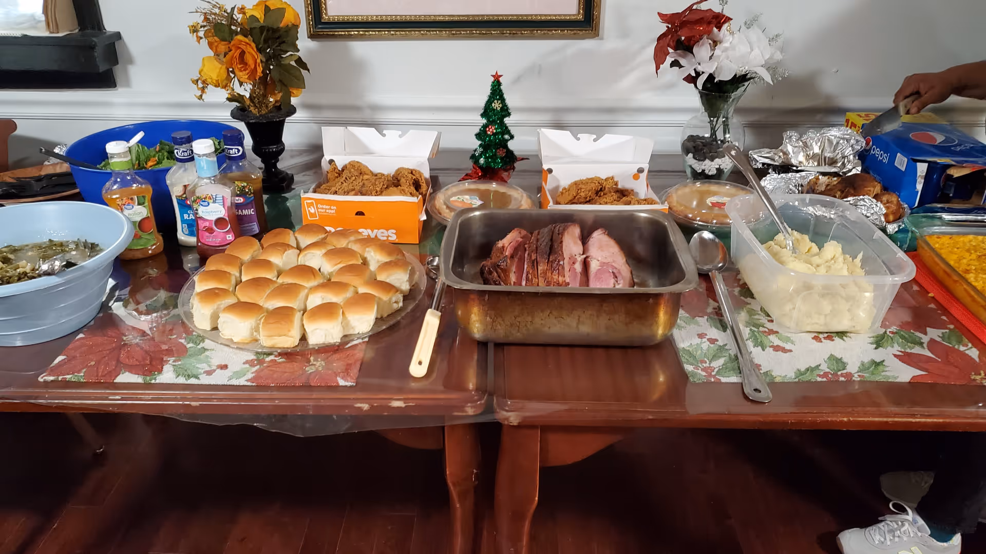 A buffet-style meal setup on two tables with various dishes including a plate of dinner rolls, a pan of sliced ham, containers of mashed potatoes, fried chicken in boxes, a bowl of greens, salad dressings, and a tray of corn. There are floral arrangements and a small Christmas tree decoration in the center. A person is seen on the right side reaching for a box of Pepsi cans.