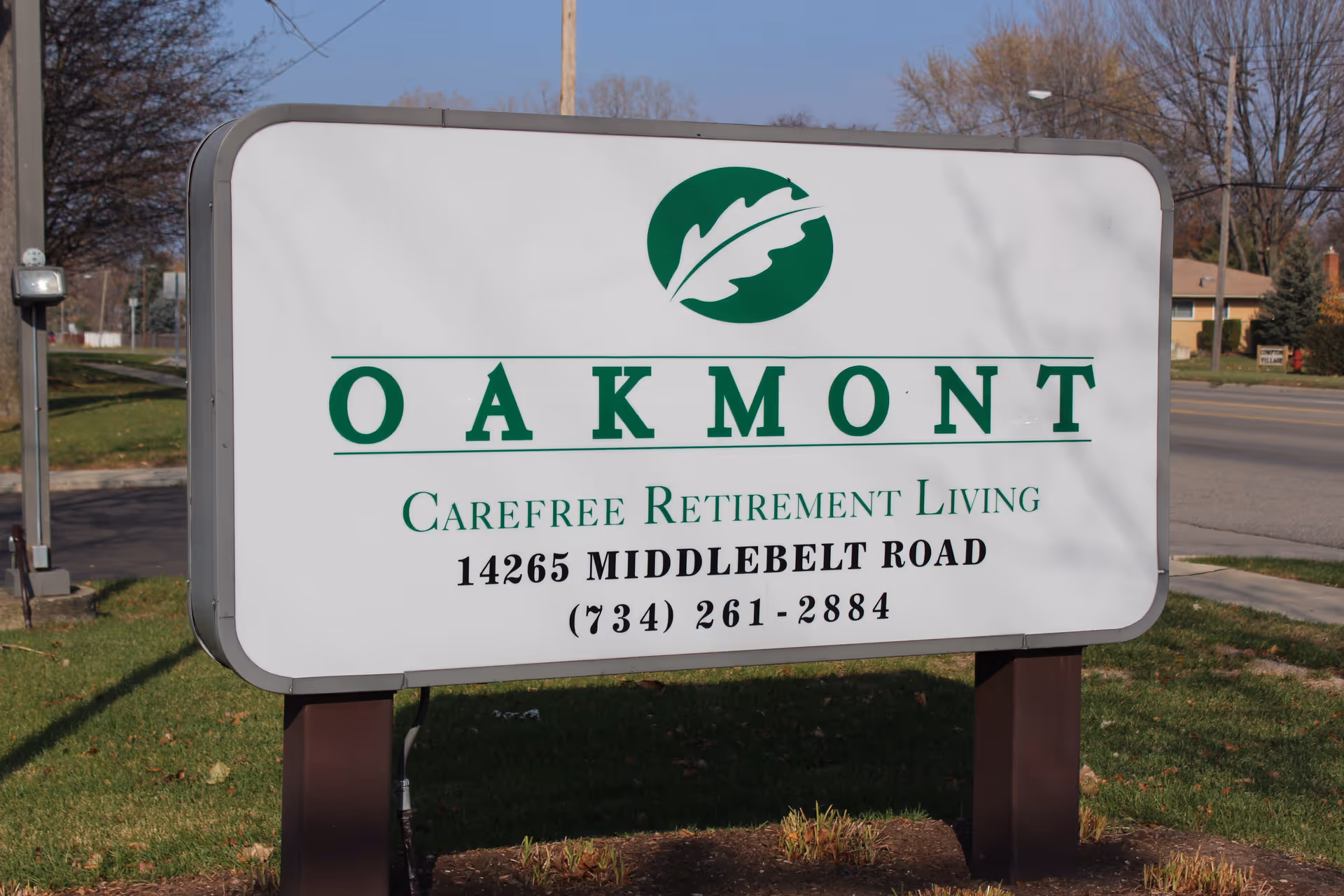 Outdoor sign for Oakmont Carefree Retirement Living located at 14265 Middlebelt Road with a phone number (734) 261-2884, set on a grassy area near a road with trees and houses in the background.