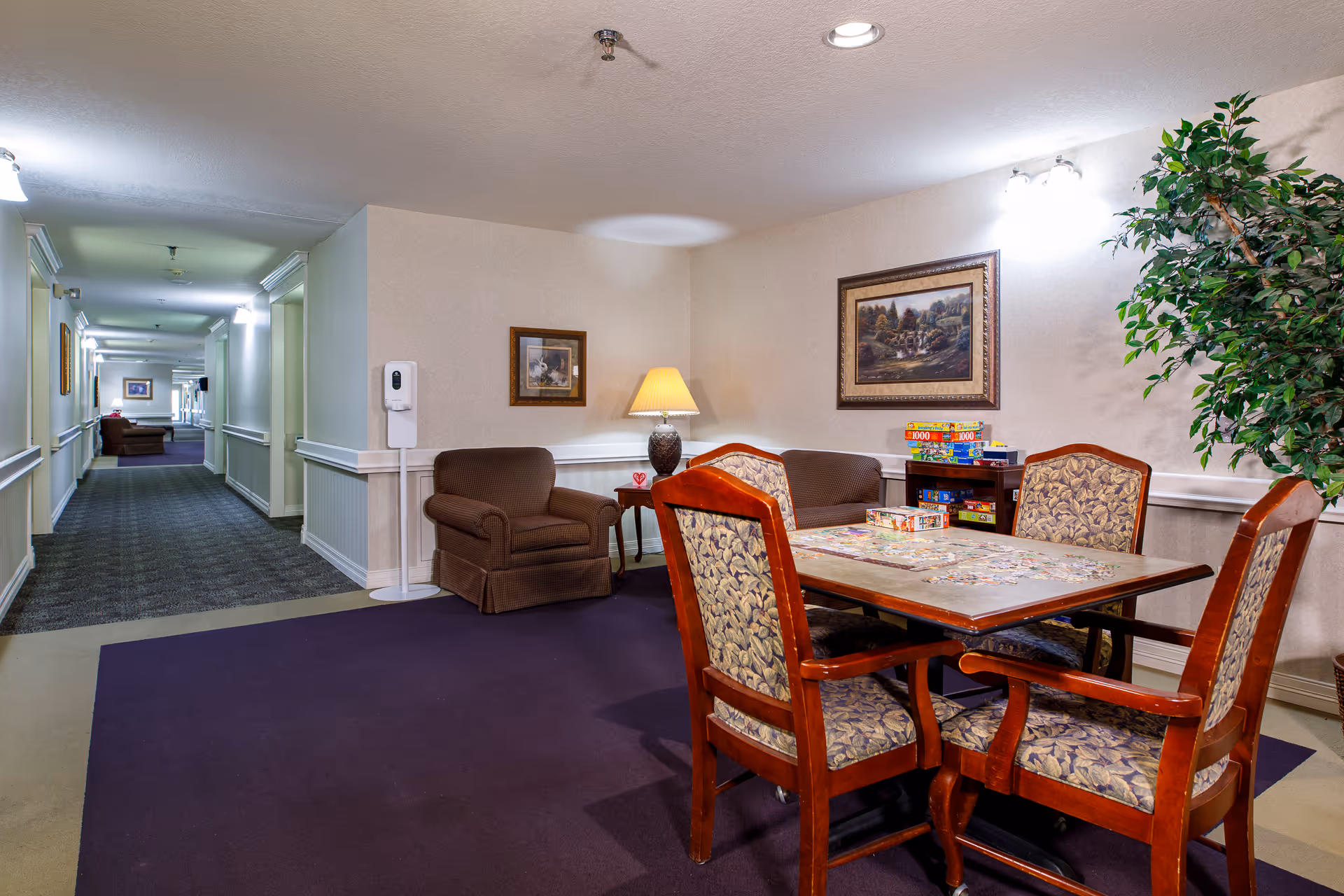 A cozy common area in an assisted living facility featuring a wooden table with four upholstered chairs arranged around it. On the table, there is a puzzle in progress. Behind the table, there is a small shelf with board games stacked on top. To the left, there is a brown armchair next to a small side table with a lamp. The walls are decorated with framed paintings, and a large artificial plant is placed in the corner. The area opens into a long, well-lit hallway with additional seating visible in the distance.