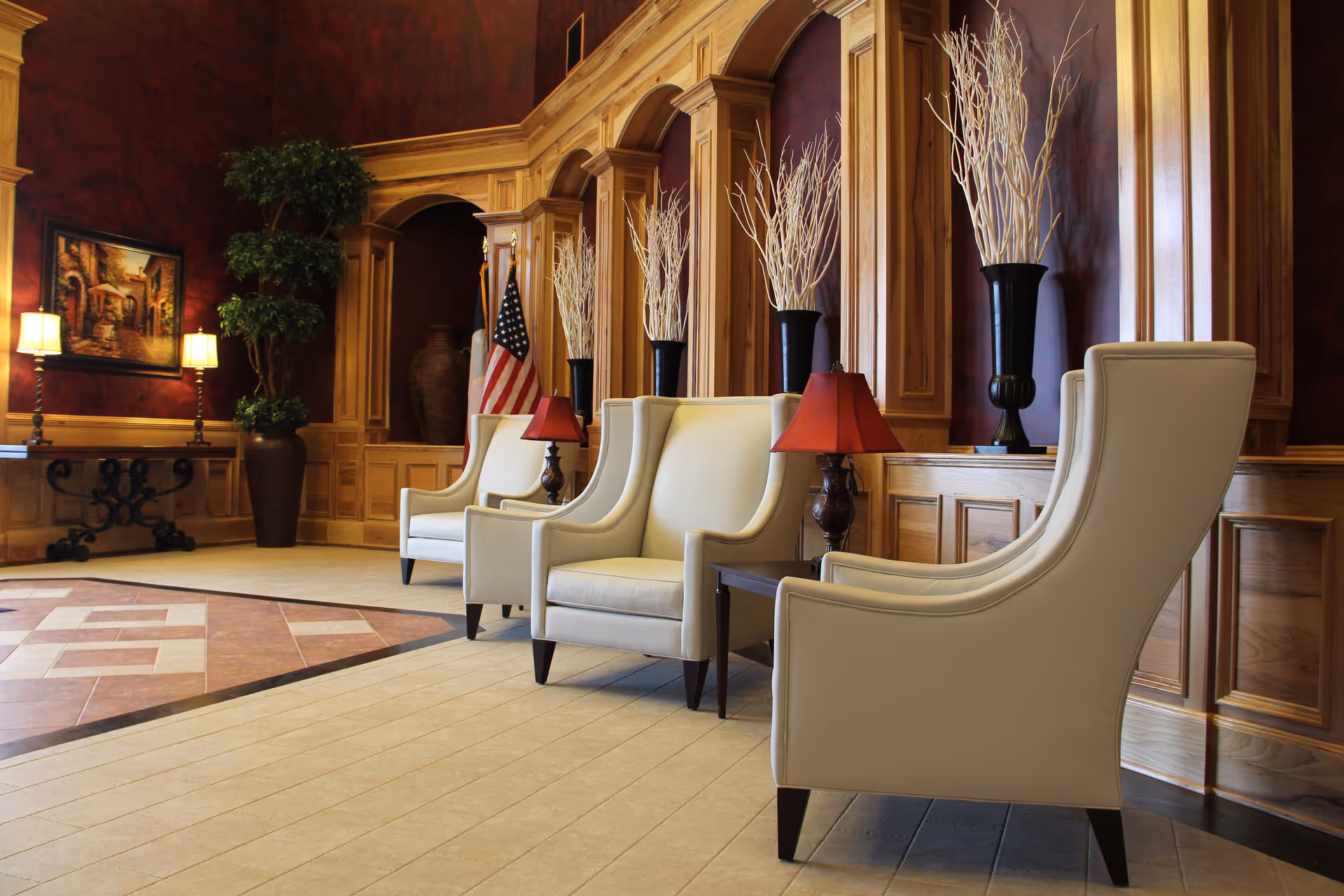 A spacious and elegant seating area with cream-colored armchairs arranged in a row against a wooden paneled wall with decorative arches. Each chair is accompanied by a small table with a red lampshade. Tall black vases with white decorative branches are placed in the wall niches. The floor features a combination of light tiles and a patterned section. In the background, there is a large potted plant, an American flag, a framed painting, and two table lamps on a console table.