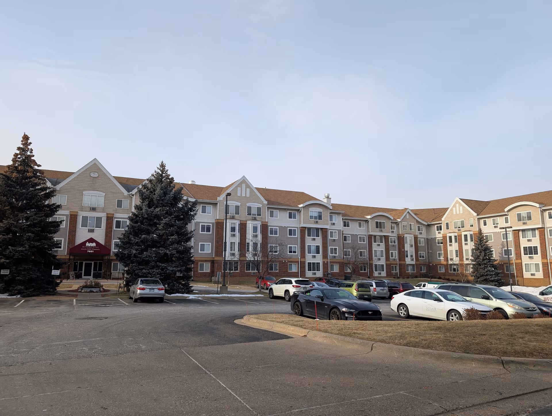 Front view of a multi-story senior living building with parked cars and trees in front under a clear sky.