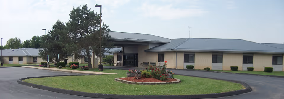Exterior view of a single-story residential care center building with a metal roof, surrounded by a paved driveway and landscaped with grass, trees, and flower beds.