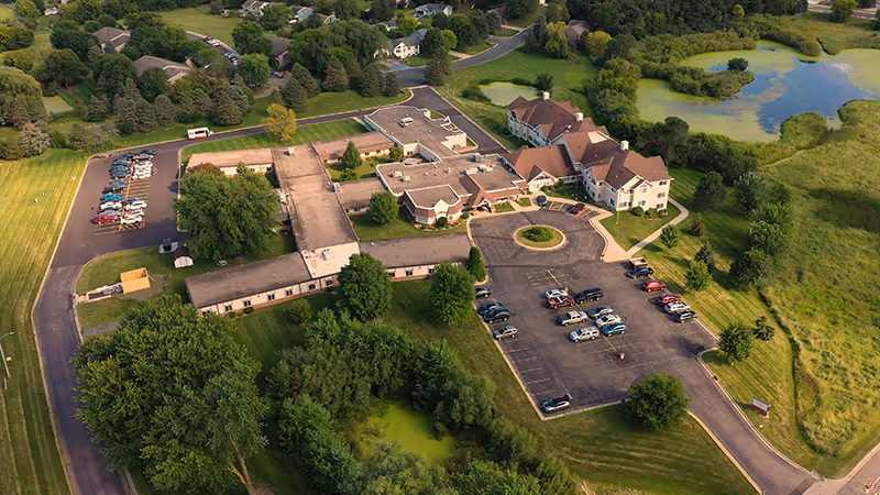 Aerial view of Mala Strana Assisted Living facility showing a large building complex surrounded by green lawns, trees, a pond, and parking lots with several cars parked. Residential houses and more greenery are visible in the background.