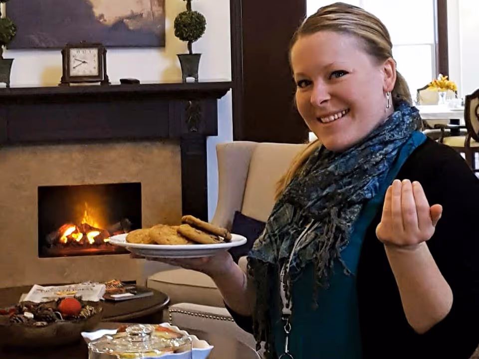 A smiling woman holding a plate of cookies in a cozy living room with a lit fireplace, a beige armchair, and a table with snacks and magazines.