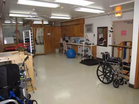 Interior of Pinehurst Nursing and Rehabilitation therapy area with wheelchair, exercise equipment, and cabinets.