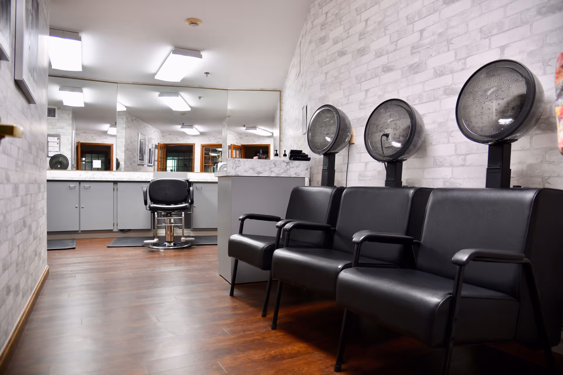 Interior view of a salon area in a senior living facility with three black salon hair dryers mounted on the wall above a row of black chairs. Across the room, there is a styling chair in front of a large mirror with cabinets underneath the countertop. The floor is wood, and the walls have a light gray brick pattern.