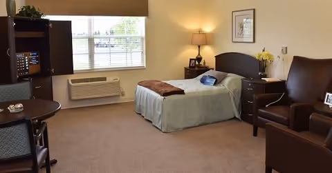 A well-lit bedroom in a senior living facility with a single bed covered in light green bedding and a brown throw blanket. Next to the bed is a bedside table with a lamp and a vase of yellow flowers. There is a comfortable brown armchair and a small table with a chair near a window with blinds. A wall-mounted air conditioning unit is below the window, and a framed picture hangs on the wall above the bed.