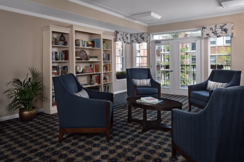 A cozy sitting area with four dark blue armchairs arranged around a dark wooden oval coffee table with books on it. Behind the chairs is a large white bookshelf filled with books and decorative items. The room has large windows and a glass door letting in natural light, with floral valances above the windows. A potted plant is placed in the corner on the left side of the room.