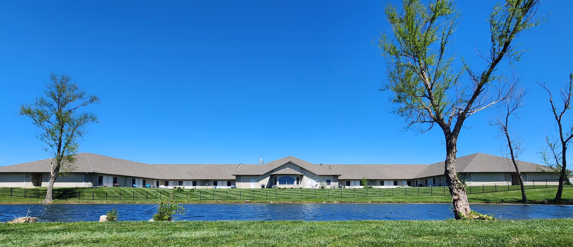 Wide exterior view of Oak Pointe Assisted Living building with a clear blue sky, a small pond in the foreground, green grass, and several trees around the pond.